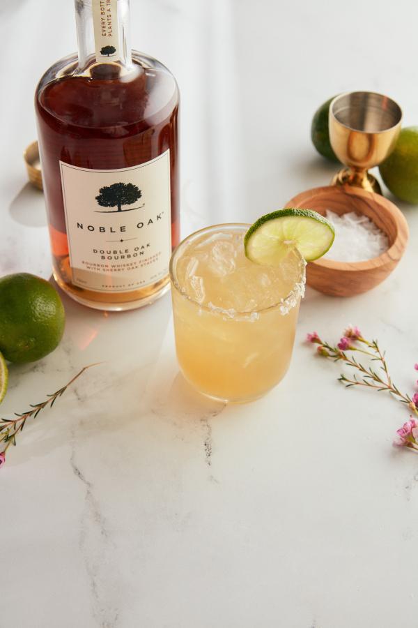 Image shows a margarita made with bourbon and a bottle of noble oak bourbon on a white marble counter top with salt, a jigger, and limes in the background.