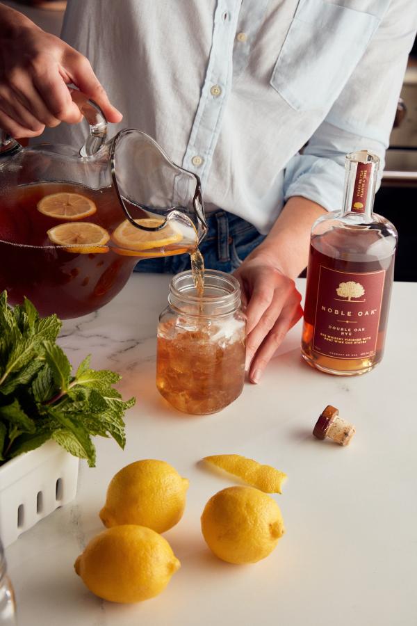 This image shows someone in a light blue button down pouring the tea into a mason jar glass. There is a bottle of noble oak rye next to her and lemons in the foreground. 