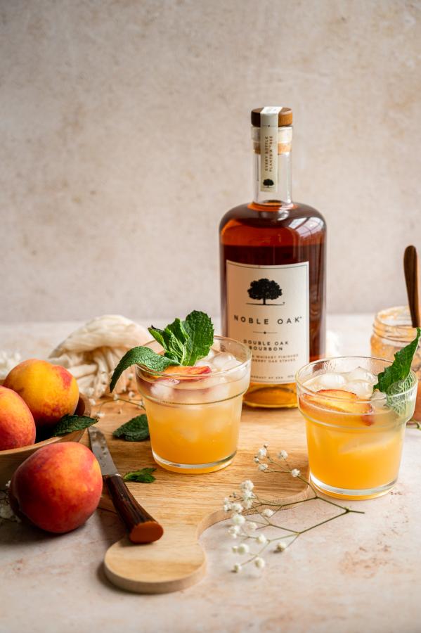 image shows two bourbon peach smash cocktails on a cutting board with peaches and a bottle of bourbon.