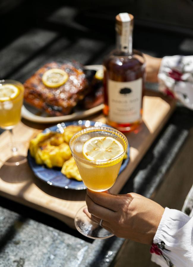 Image shows a cocktail garnished with grilled lemon with a spread of barbecue and a bottle of noble oak in the background. 